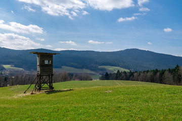 J&auml;gersitz auf dem Feld mit B&auml;umen und Wald und Bergen im Hintergrund sowie Wolken am Himmel im Bayerischen Wald