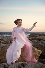 Fototapeta na wymiar capricious restless princess girl sitting on a stone by the sea in a pink dress with veil and crown