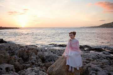 pensive sad princess girl in a pink dress and a diadem is sitting on a rock on the ocean seashore...