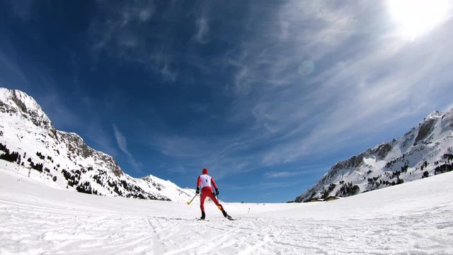 slow motion one male cross country skier in race suit with start number skating on cross country ski run in mountains on sunny winter day in 4K rear view
