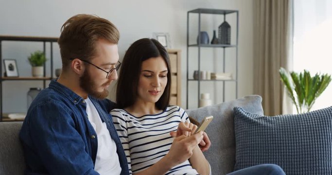 Attractive young just married couple sitting on the sofa in the living room and watching something on the smartphone device while woman taping on it. Indoors