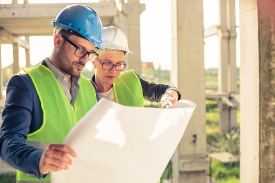 Young female and male engineers or business partners at construction site, working together on building's blueprint. Architecture and teamwork concept.