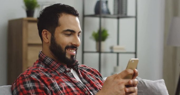 Close up of the attractive Middle Eastern man in the motley red shirt typing and chatting on the smartphone at home. Inside