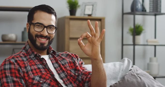 Portrait of the attractive Middle Eastern man in the glasses and motley red shirt smiling to the camera and showing ok gesture in the living room. Close up. Inside
