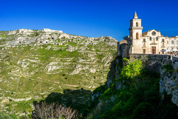 Fototapeta na wymiar Horizontal View of the Church of Peter and Paul Saints on the Background of the Caves of the Gravina of Matera on Blue Sky Background. Matera, South of Italy