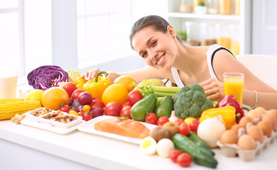 happy young housewife sitting in the kitchen preparing food from a pile of diverse fresh organic fruits and vegetables