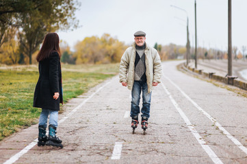 Happy old man goes on rollers with his daughter on the road in autumn park. Happy and active pensioner.