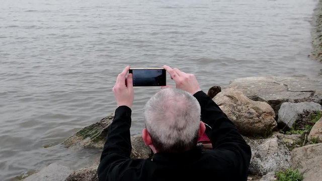 Midle aged man sitting on the shore of the lake. Solitude mature man recording video by mobile phone on the bank. Concept of loneliness