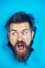 Close up portrait of surprised man looking through hole in blue paper. Bearded man making hole in paper. Shocked man with beard and mustache looking through hole in blue paper.