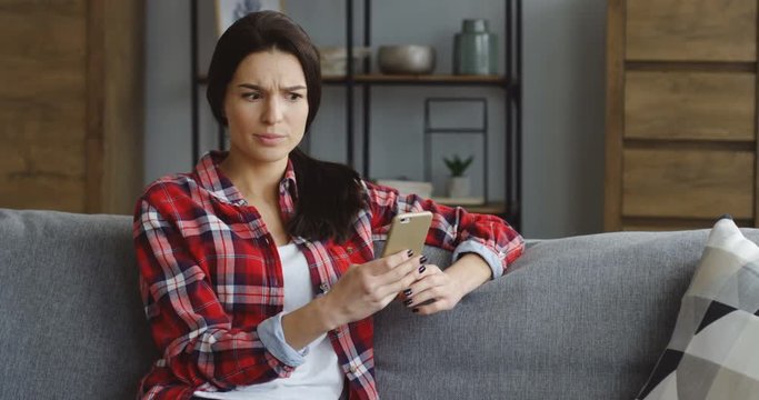 Pretty brunette young woman in the red motley shirt reading something on the smartphone and getting to know shoking news at home. Indoors