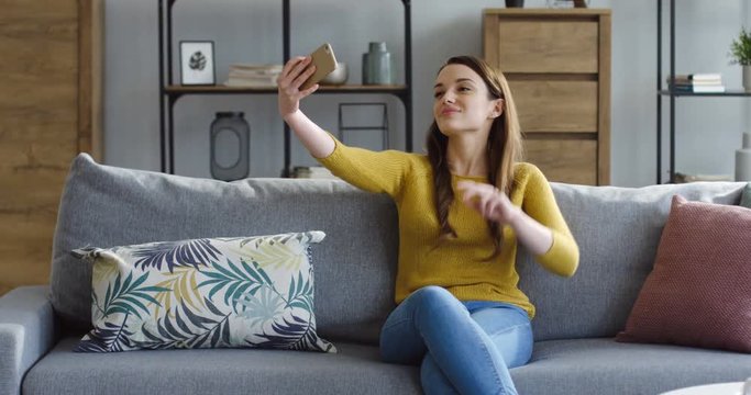 Brunette beautiful woman in the yellow sweater and jeans making nice selfies on the smartphone while sitting on the gray couch in the cozy room. Indoors