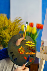 Artist in blue sweater draws mixes oil paints with palette knife close-up. He holds palette and brush bristle. Flowers in the background