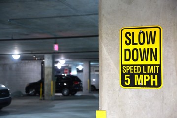 Yellow and Black Slow Down Speed Limit 5 Mph Sign in a Parking Garage with Cars Out of Focus in the Background © kthx1138