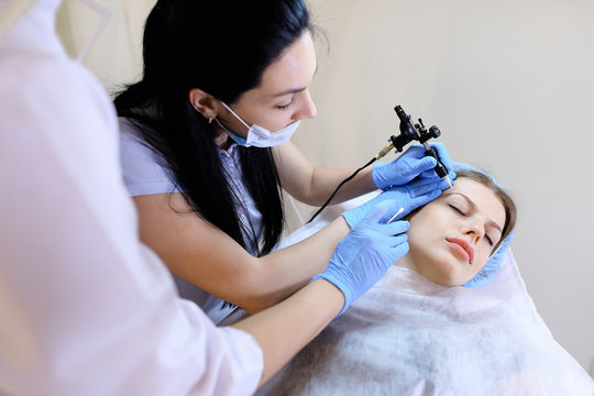 Female cosmetologist making tattooing procedure for woman by machine. Concept of beauty industry and tattooing eyesbrows.
