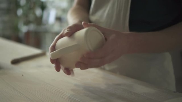 Close up of mug, which is being checked before painting in ceramic studio. Male ceramist in dickey is standing at the wooden table, holding cup in his hands, twisting it and touching the surface by