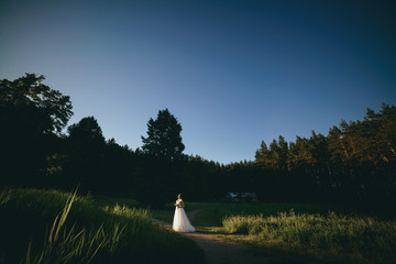 Obraz premium Young woman in a white dress is standing on the field. Portrait of girl outdoor. Romantic young woman posing on the background of sunset sky.