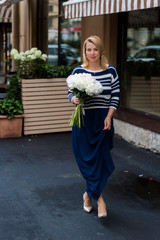 Fototapeta premium Young blonde woman in blue dress with peonies bouquet outdoors at city street