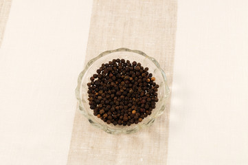 Black peppercorn on a round glass plate on a striped linen cloth tablecloth.