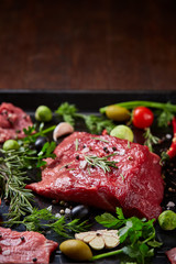 Flat lay of raw beefsteak with vegetables, herbs and spicies on metal tray, close-up, selective focus