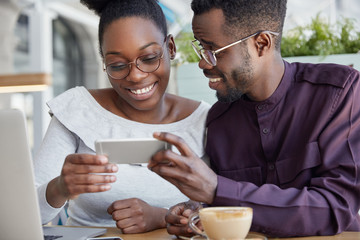 Cropped shot of deighted happy African couple holds smart phone horizontally, watch interesting video, have coffee break, smile joyfully, wears round spectacles. Dark skinned woman and man friends