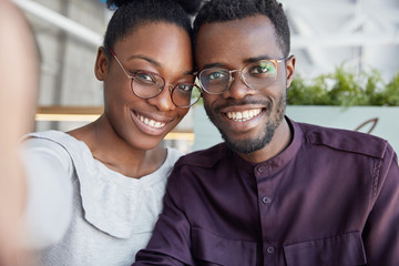 Fototapeta premium Happy dark skinned young male and woman bloggers make picture for profile, pose for selfie, share photos in social network accounts, wears spectacles, have broad smiles and white perfect teeth