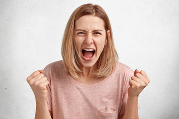 People and aggression concept. Irritated young female model with bobbed hairdo, dressed in casual clothing, clenches fists in anger, has quarrel with husband, poses in studio againt white background