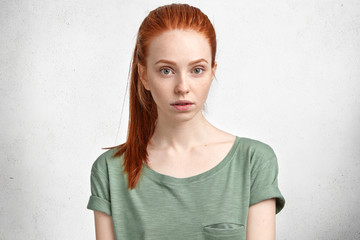 Portrait of ginger lovely woman with freckled skin and mysterious look, dressed in casual t shirt, isolated over white concrete background. Redhead young female student rests after lectures.