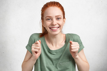 Overjoyed red haired beautiful happy young woman celebrates winning contest, dressed in casual t shirt, has broad smile, isolated over white concrete background. People, happiness and emotions