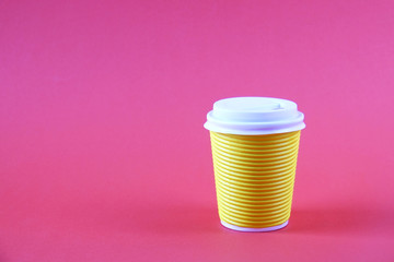 Bright yellow disposable safe heat resistant double walled paper take out cup of coffee to go. Colorful eco-friendly cardboard mug for hot beverages, whitecap on pink background, close up, copy space.