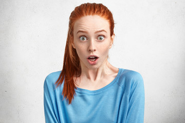 Portrait of shocked bugged eyed pretty redhead young woman expresses great surprisment, hears unexpected news, doesn`t expect to meet somebody, wears blue sweater, isolated over white background