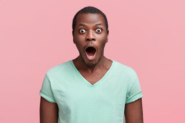 Portrait of dark skinned male feels astound, finds out about wreck, keeps jaw dropped out and eyes bugged, dressed in casual t shirt, isolated over pink background. Impressed African American man