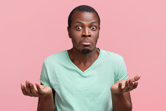 Horizontal shot of hesitant handsome dark skinned male shrugs shoulders, being uncertain and unsure, curves lower lip and stares, poses against pink background. Confused man has some doubts.