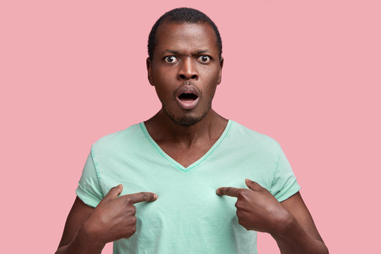 Displeased annoyed African American male model indicates at t shirt for your design or logo, frowns face and being dissatisfied with something, isolated over pink background. Negative emotions