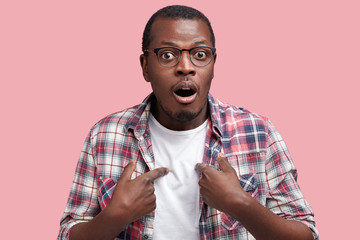 Photo of embarrassed African American male indicates at himself with puzzled expression, being shocked to be chosen to participate in competition, hesitates about something isolated on pink wall