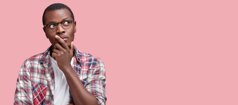 Thoughtful African American male being deep in thoughts keeps hand on mouth, wears casual checkered shirt and spectacles, contemplates about something, poses against pink studio background, copy space