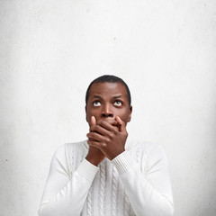 Vertical shot of terrified serious dark skinned male covers mouth with hands, looks with shocked expression up, notices something awful, poses against white concrete background with copy space