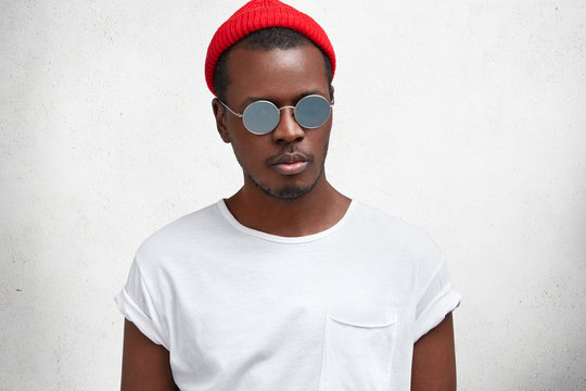 Portrait of fashionable hipster guy in trendy eyewear, red hat and casual white t shirt, gong to meet with friends, has serious confident look, poses against concrete wall. People, ethnicity, style