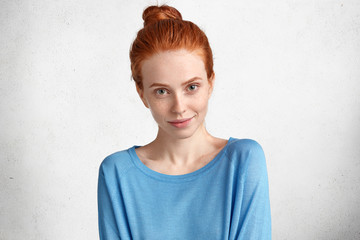 Headshot of attractive young red haired female with hair knot, has pleased look, dressed in casual blue sweater, going to meet with best friend after work, isolated over concrete background.