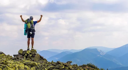 Foto auf Acrylglas Mann auf dem Gipfel des Berges. Emotionale Szene. Junger Mann mit Rucksack, der mit erhobenen Händen auf einem Berg steht und Bergblick genießt. Wanderer auf der Bergspitze. Sport- und aktives Lebenskonzept. © bilanol