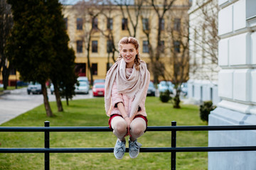 Thoughtful pretty female student girl with two braids sitting on the handrails over university building background. Front view, full height.