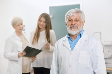 Confident old doctor staying in front of assistant and patient. Looking expirienced, skilled,...