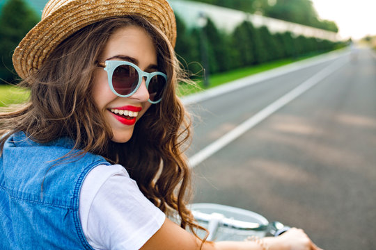 Closeup portrait of cute girl with long curly hair in hat  driving a bike on road. She wears  jerkin, blue sunglasses. She is smiling to camera, view from back.