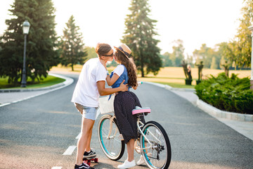 Young couple in love on road on sunset. Pretty girl with long curly hair in hat and long skirt holds a bike, handsome guy on skateboard hugging and going to kiss her.