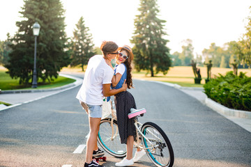 Young couple of romantic people on road on sunset. Pretty girl with long curly hair in hat and long skirt holds a bike, handsome guy on skateboard hugging and looking at her.