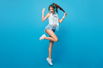 Fototapeta na wymiar Active girl with long curly hair in tail in jump on blue background in studio. She wears white T-shirt, shorts. She is listening to music with blue headphones.
