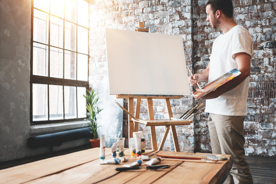 Man Artist Stands In Front Of A Blank Canvas On Easel With Palette In Loft Art Studio. Male Painter Hold Paintbrush In Hand And Mix Paint. Hobby Concept