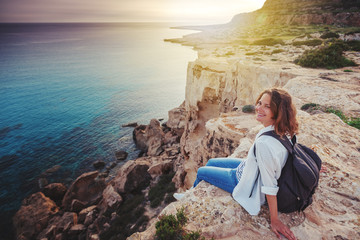 A stylish young woman traveler watches a beautiful sunset on the rocks on the beach, Cyprus, Cape Greco, a popular destination for summer travel in Europe