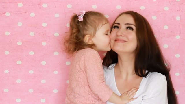 Mother and daughter embrace, smiles, have fun, laugh and kissed. Close-up portrait young woman and her child on pink background.