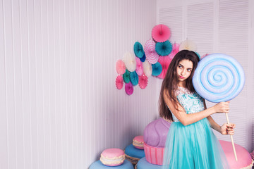 beautiful girl in blue dress hold in hand huge candy in studio with decor of macaroons. Copy space.