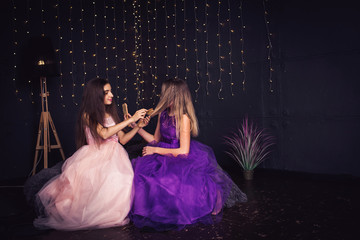 Cheerful girlfriends. Two girls comb each other's hair, sitting side by side in studio on dark...
